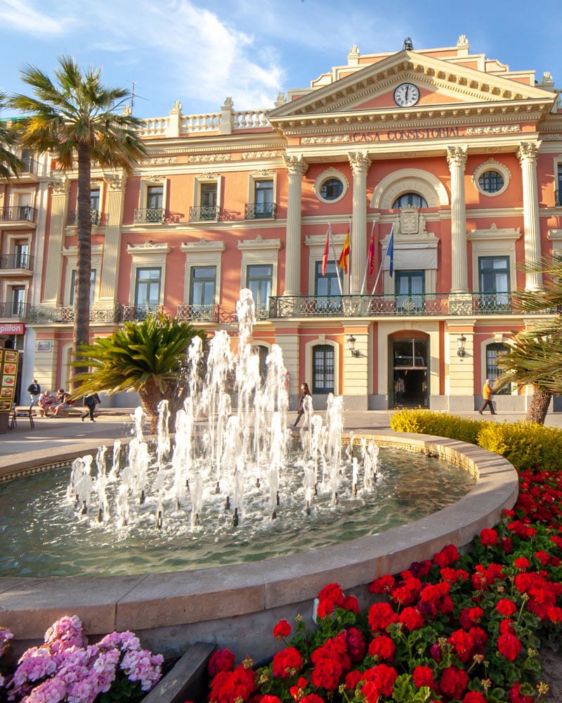 A pink building and a water fountain with palm trees in Murcia Spain