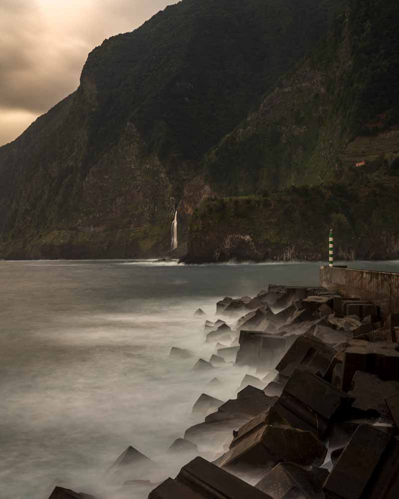 Long exposure of waves crashing against a row of rocks, while a waterfall tumbles into the ocean beyond